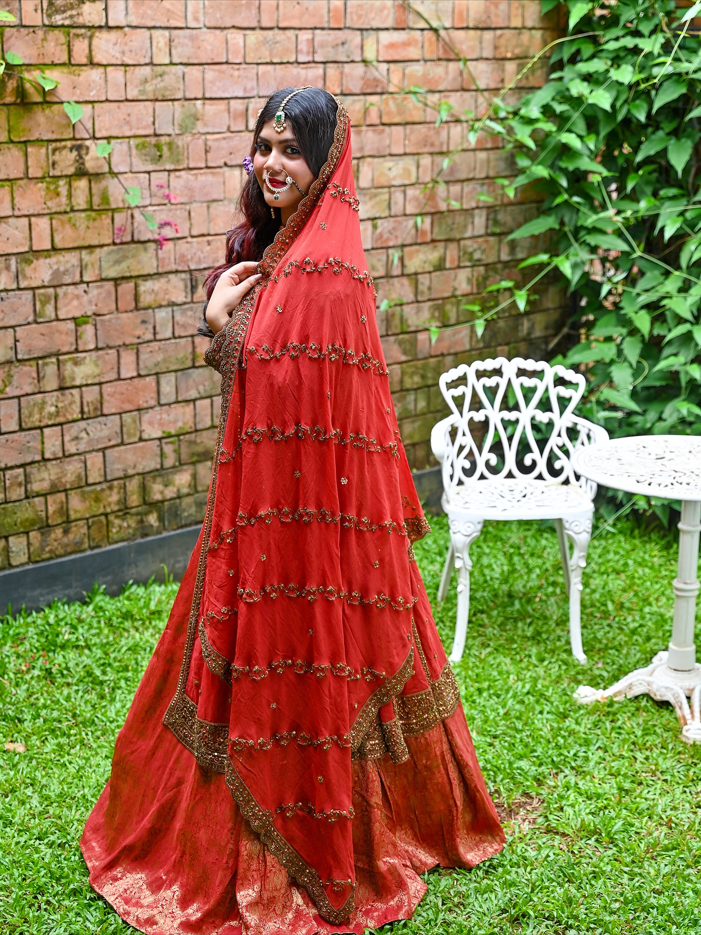 A bride in deep crimson velvet dupatta with gold embroidery, posed by a brick wall and Victorian iron table