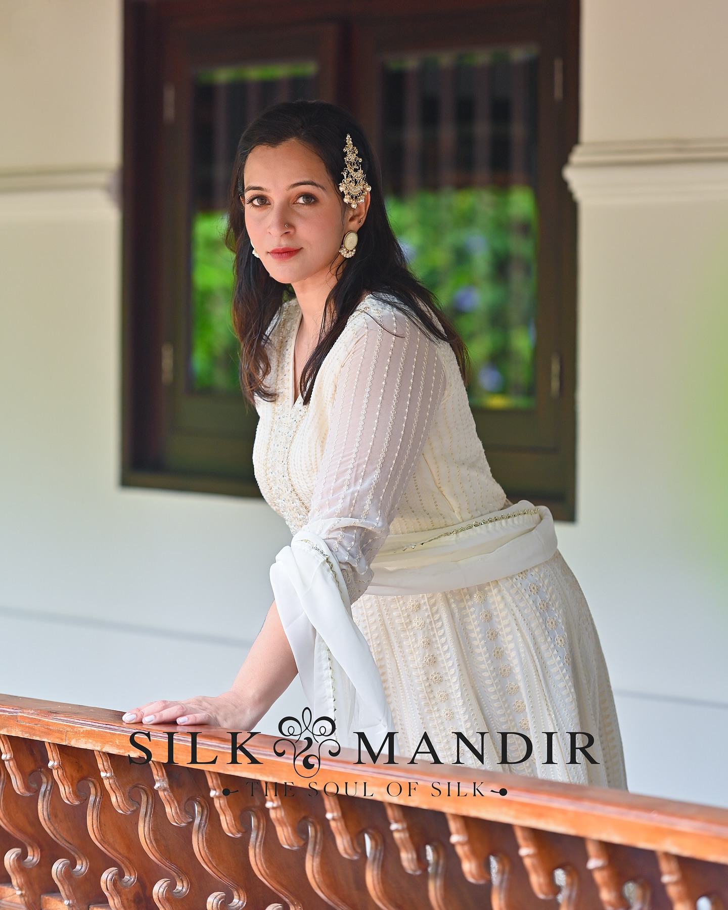 A woman in pearl-embroidered cream Anarkali leaning on a carved wood balcony of a Kerala heritage home
