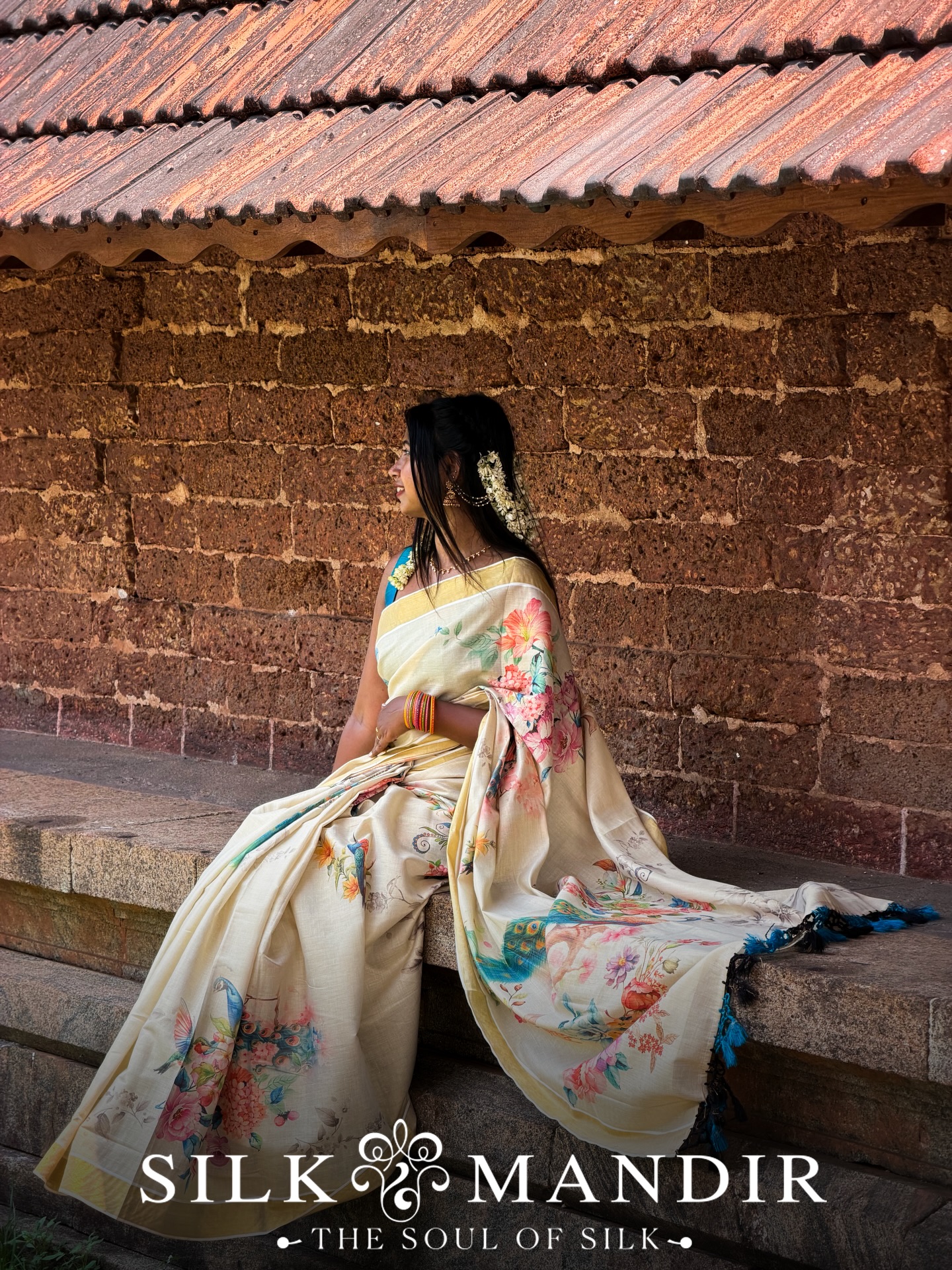 A young woman in cream Kerala kasavu saree seated on temple steps with a laterite wall behind her